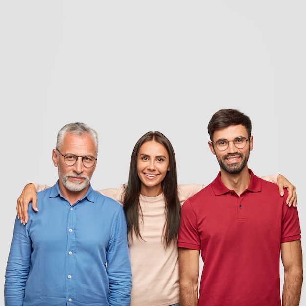 Posa della famiglia felice per fare foto comuni: padre senior positivo, figlia adulta e figlio si abbracciano, sorriso amichevole, posa contro il muro bianco. Concetto di persone, generazione e relazioni