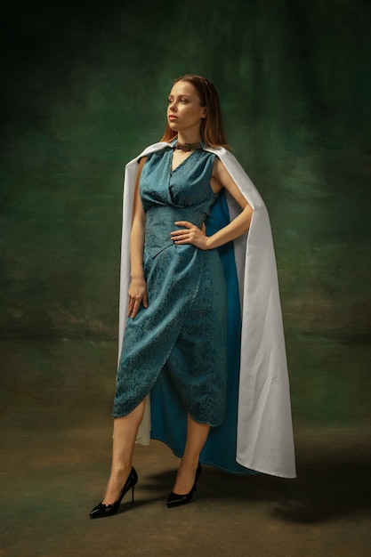 Posa di eleganza. Ritratto di giovane donna medievale in abiti vintage blu su sfondo scuro. Modello femminile come duchessa, persona reale. Concetto di confronto di epoche, moderno, moda, bellezza.