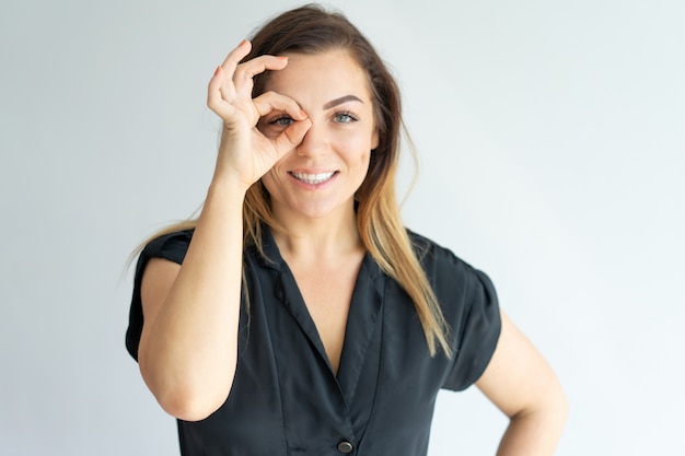 Positiva attraente giovane donna facendo segno ok e guardando attraverso le dita alla macchina fotografica.