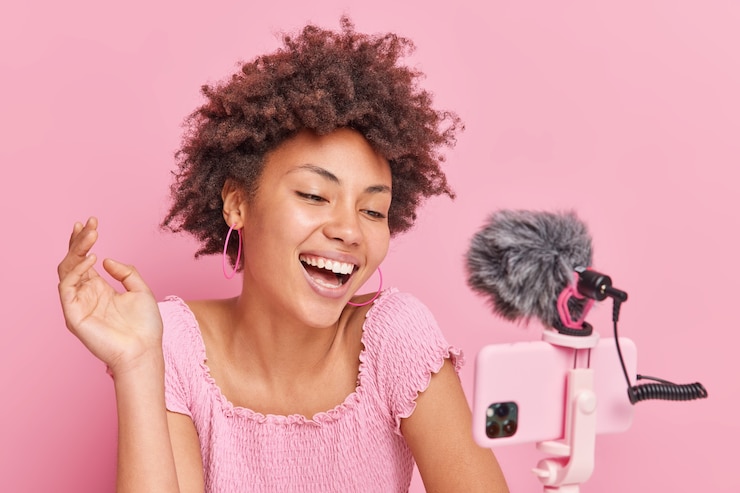 Foto gratuito positiva bella bruna blogger afroamericana focalizzata sullo smartphone sul treppiede fa sì che lo streaming online abbia il proprio canale sorrisi posa felicemente contro il muro rosa. concetto di vlogging.