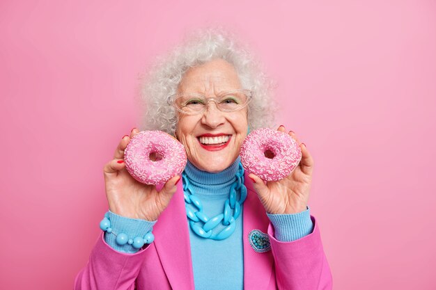 Positiva bella donna europea anziana rugosa tiene due deliziose ciambelle glassate sorrisi ha ampiamente buon umore applica trucco vestiti e gioielli alla moda