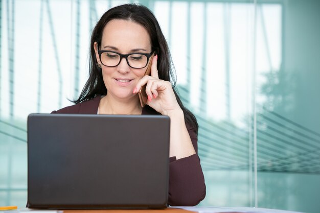 Positiva bella imprenditrice in bicchieri parlando al telefono cellulare, lavorando al computer in ufficio, utilizzando il computer portatile al tavolo