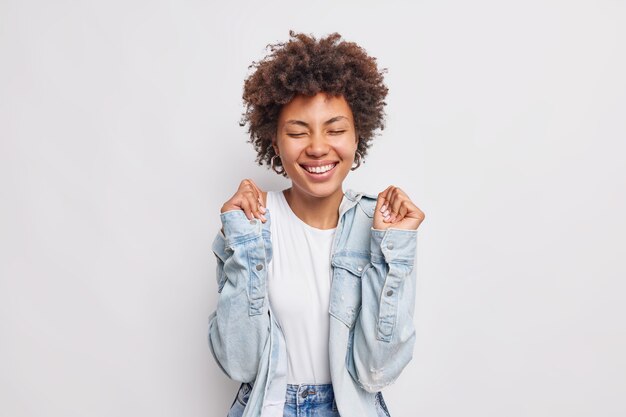 Positiva giovane donna dai capelli ricci alza le mani in attesa di risultati gioisce notizie fantastiche tiene gli occhi chiusi sorrisi indossa ampiamente la camicia di jeans isolata sul muro bianco