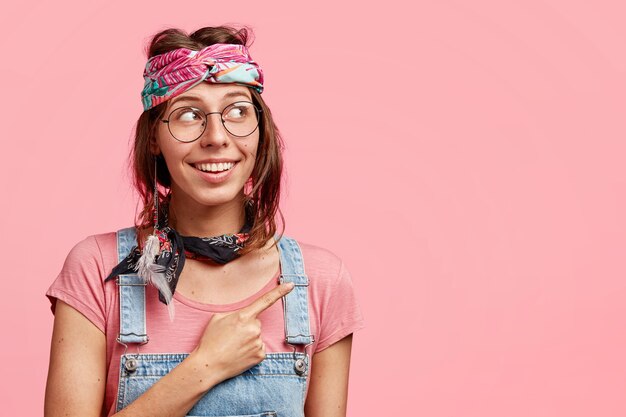 Positiva giovane donna hippy con espressione gioiosa punti da parte, vestita con fascia alla moda e tuta di jeans