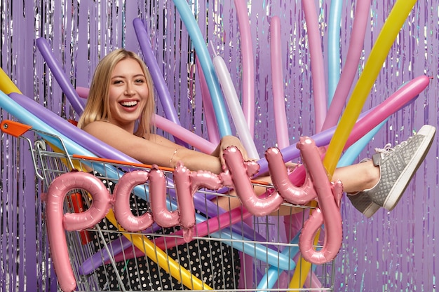 Positiva la donna caucasica con i capelli chiari, pone nel carrello con palloncini colorati di modellazione, essendo di buon umore, celebra l'anniversario, isolato sopra il muro viola