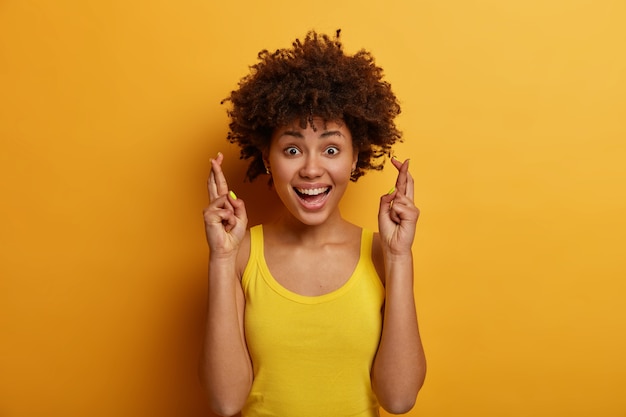 Positiva ragazza carina con acconciatura afro crede che i sogni diventino realtà, tiene le dita incrociate, aspetta che sia successo qualcosa di buono, vestita casualmente, ride e guarda direttamente, posa al coperto