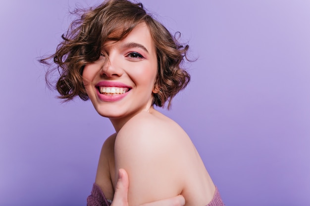 Positiva ragazza con capelli castani lucidi che gode del servizio fotografico Ritratto dell'interno della signora allegra con taglio di capelli corto isolato sulla parete viola.