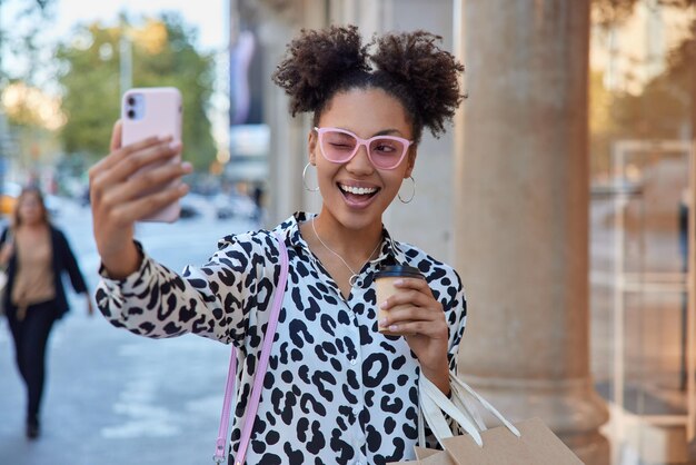 Positiva ragazza spensierata con due chignon strizza l'occhio alle pose degli occhi alla fotocamera dello smartphone fa selfie bevande caffè aromatico tiene borse della spesa ha buon umore passeggiate in strada in città.