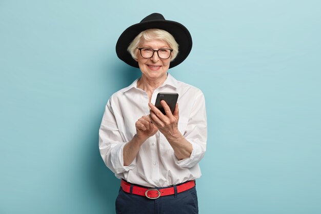 Positiva signora anziana con la faccia rugosa, felice finalmente di imparare a usare smartphone e internet, indossa occhiali trasparenti, cappello nero, camicia e pantaloni eleganti, isolati sopra il muro blu.