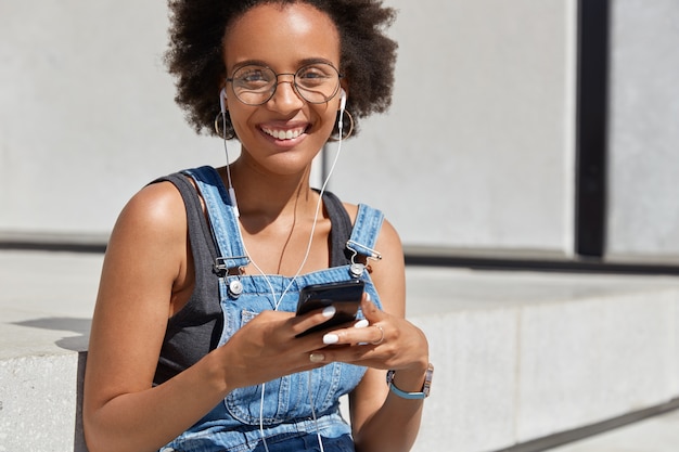 Positiva signora dalla pelle scura con un sorriso a trentadue denti, ascolta l'audio al cellulare digitale, è felice di ascoltare la canzone preferita, gode del suono puro, indossa occhiali rotondi e abiti casual, posa all'esterno