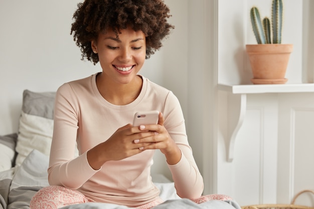 Positiva signora nera con taglio di capelli afro, tiene il cellulare moderno, riconsidera il messaggio di testo ricevuto da un amico, digita il feedback, vestita in camicia da notte, si siede da solo su un comodo letto, ha una giornata pigra