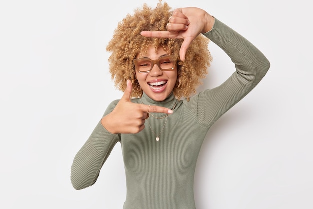 Positiva spensierata giovane donna allegra con i capelli ricci guarda attraverso la cornice della mano foto momento essendo creativo e felice indossa occhiali dolcevita isolato su sfondo bianco scatta foto