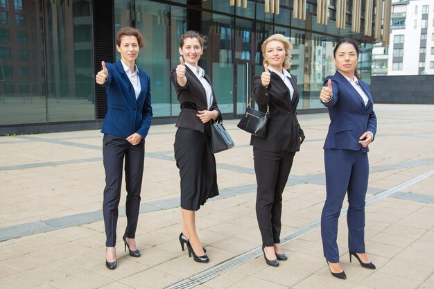 Positiva squadra di donne d'affari fiducioso in piedi insieme vicino all'edificio per uffici, mostrando come il gesto, che fa il pollice in su, guardando la fotocamera. Lunghezza intera. Il lavoro di squadra e il concetto di successo aziendale