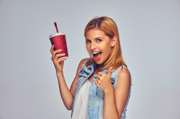 Positivo e la femmina bionda sorridente tiene una tazza di carta rossa con succo isolato su sfondo grigio.