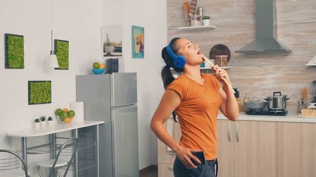 Positivo giovane donna che canta sul cucchiaio di legno al mattino. Casalinga energica, positiva, felice, divertente e carina che balla da sola in casa. Divertimento e svago da soli a casa