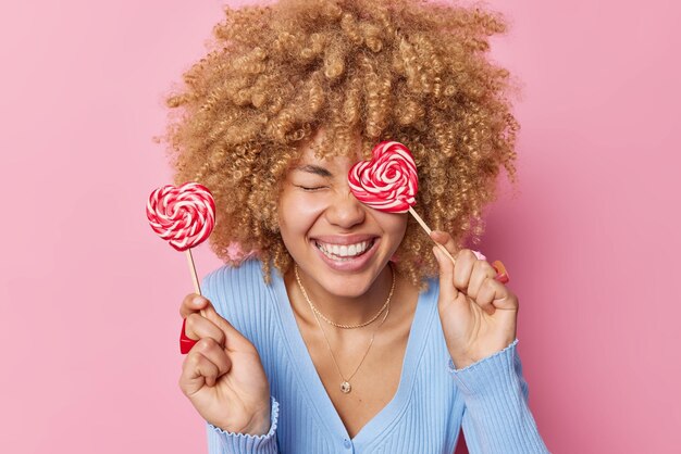 Positivo giovane donna con capelli ricci biondi hlds due caramelle a forma di cuore caramello mantiene gli occhi chiusi sorrisi a trentadue denti indossa un maglione blu casual isolato su sfondo roseo Concetto di dente dolce