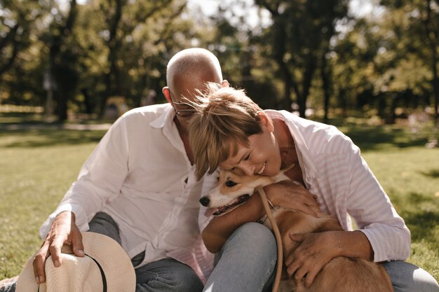 Positivo laici con capelli biondi in camicia a righe sorridente, abbracciando corgi e in posa con l'uomo in camicia bianca moderna nel parco.