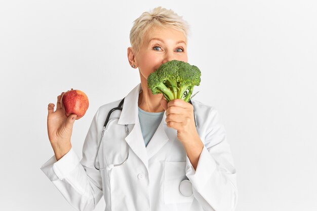 Positivo medico donna di mezza età che tiene mela e broccoli, raccomandando una dieta a base vegetale. Dottoressa divertente che suggerisce di mangiare verdure che forniscono nutrienti vitali, a basso contenuto di grassi e calorie