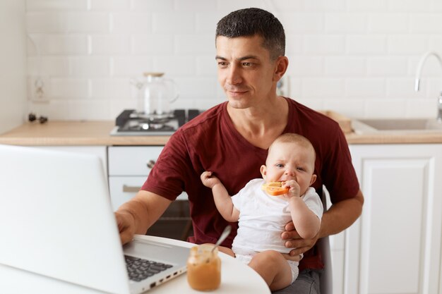 Positivo ottimista bel libero professionista maschio che indossa la maglietta bordeaux, posa in cucina bianca, seduto davanti al computer portatile con il bambino in mano e lavora online.