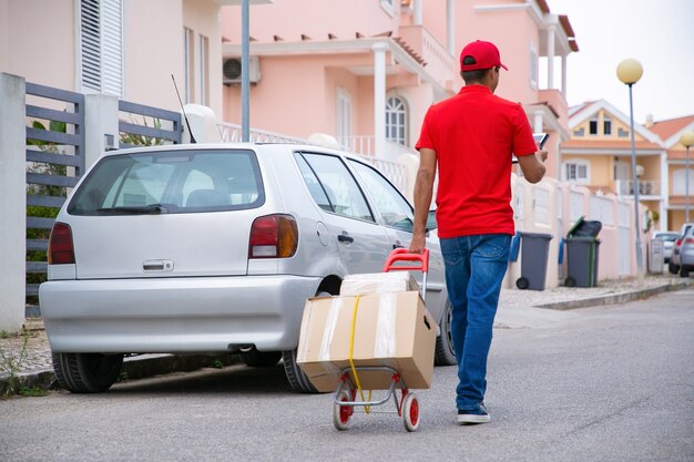 Postino che tiene tablet e carrello con ruote con scatole di cartone. Corriere professionale caucasico in uniforme rossa che cammina sulla strada con i pacchi di cartone sul carrello. Servizio di consegna e concetto di posta