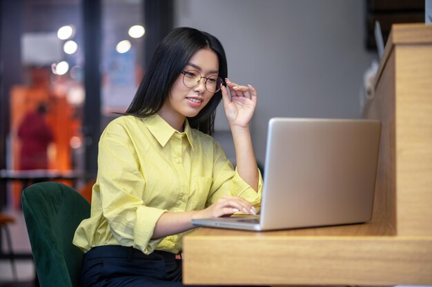 Posto di lavoro. Giovane donna asiatica sveglia in occhiali al computer portatile che mangia caffè