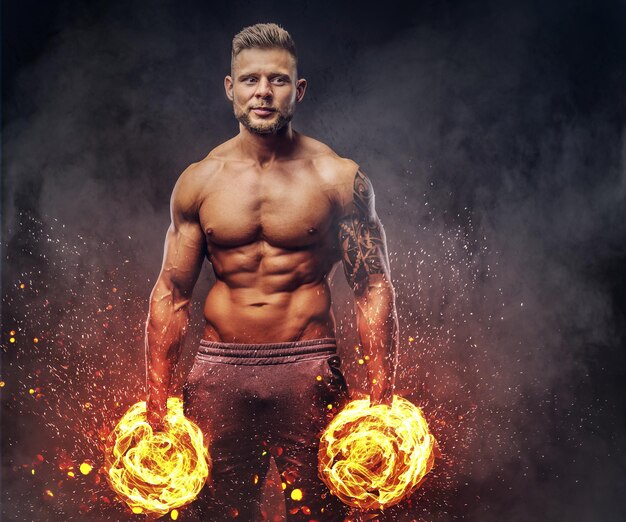 Potente bodybuilder elegante con tatuaggio sul braccio, in posa con manubri in uno studio. Concetto di arte del fuoco.