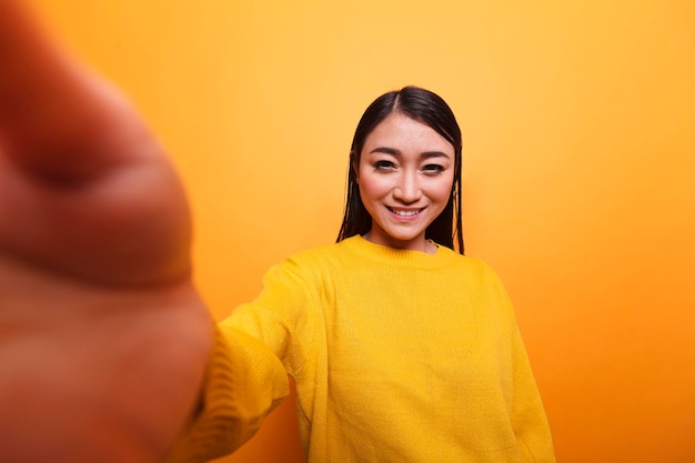 POV di un influencer sorridente felice che indossa un maglione giallo che si fa selfie con la fotocamera mentre è su sfondo arancione. Vlogger spensierato abbastanza sicuro che sorride alla telecamera mentre registra un vlog quotidiano.