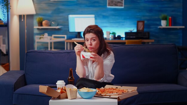 POV di una giovane donna che mangia cibo cinese con le bacchette mentre si guarda la televisione dopo il lavoro in soggiorno. Adulto che mangia fast food sul tavolo godendo di un pasto asiatico e guardando la fotocamera
