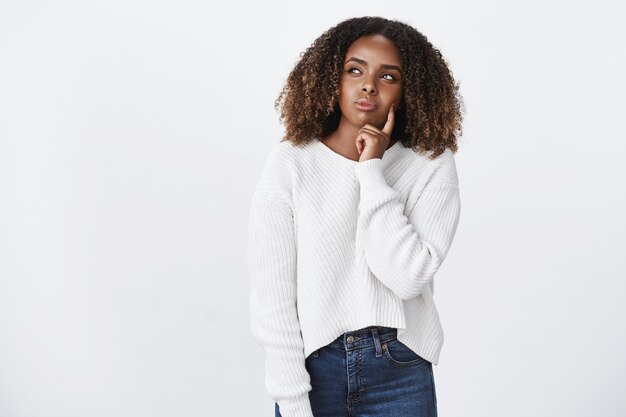 Premurosa attraente giovane donna afroamericana con i capelli ricci in maglione decidendo cosa indossare in piedi sul muro bianco strizzando gli occhi come pensare, toccando la guancia