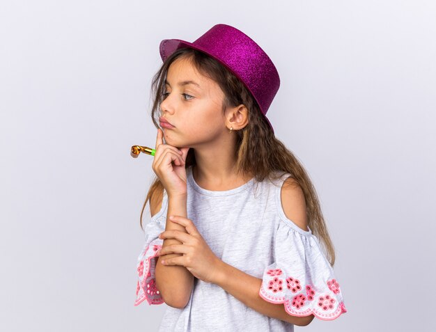 premurosa bambina caucasica con cappello da festa viola che mette la mano sul mento tenendo il fischio della festa e guardando il lato isolato sul muro bianco con spazio di copia