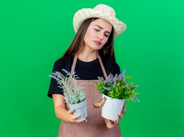 premurosa bella ragazza giardiniere in uniforme che indossa cappello da giardinaggio tenendo e guardando il fiore in vaso di fiori isolato sul verde