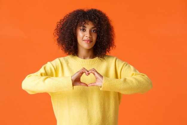 Premurosa e tenera, adorabile donna afroamericana romantica esprime i suoi sentimenti, mostra il gesto del cuore, confessa l'amore il giorno di San Valentino, in piedi il muro arancione felice.