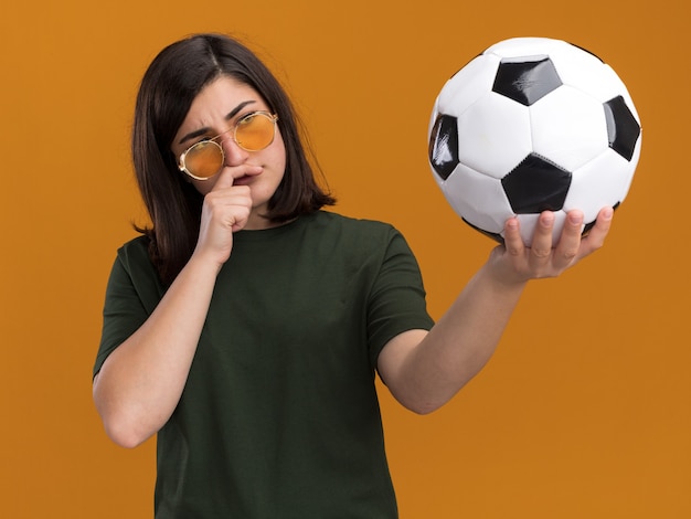 Premurosa giovane bella ragazza caucasica con gli occhiali da sole mette la mano sul mento tenendo e guardando la palla