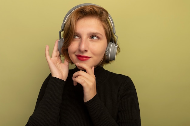 Premurosa giovane ragazza bionda che indossa le cuffie che toccano il mento e le cuffie che guardano il lato immergendosi nei sogni isolati sulla parete verde oliva con spazio di copia