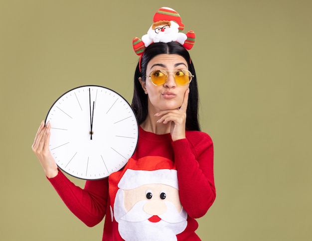 Premurosa giovane ragazza caucasica che indossa la fascia di Babbo Natale e maglione con gli occhiali tenendo l'orologio tenendo la mano sul mento guardando il lato isolato su sfondo verde oliva con spazio di copia