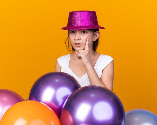 Premurosa giovane ragazza caucasica con cappello da festa viola mettendo la mano sul mento e in piedi con palloncini di elio isolati sulla parete arancione con spazio di copia