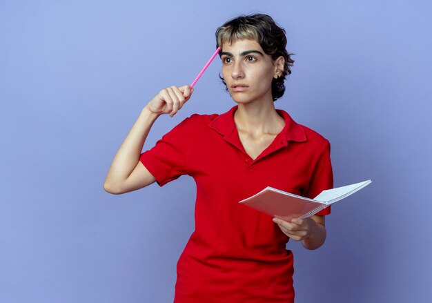 Premurosa giovane ragazza caucasica con pixie haircut holding pen e appunti toccando la fronte con la penna e guardando il lato isolato su sfondo viola con spazio di copia