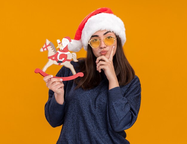 Premurosa giovane ragazza caucasica in occhiali da sole con santa hat mette il dito sul mento e tiene santa sulla decorazione del cavallo a dondolo isolato su sfondo arancione con spazio di copia