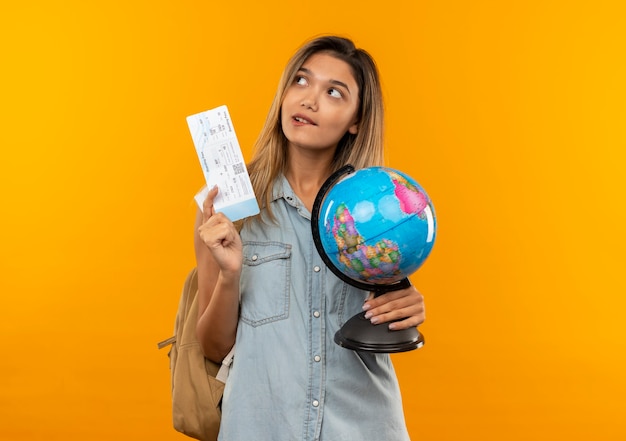 Premurosa giovane studentessa graziosa che indossa la borsa posteriore che tiene il biglietto aereo e il globo guardando il lato e mordendosi il labbro isolato sulla parete arancione