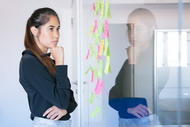 Premurosa imprenditrice latina azienda pennarello e note di lettura sulla parete di vetro. Lavoratore femminile grazioso fiducioso concentrato in vestito che pensa all'idea per il progetto.