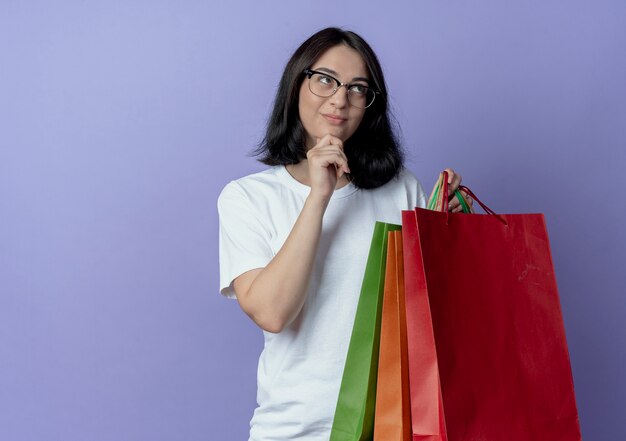 Premurosa ragazza abbastanza caucasica giovane azienda borse della spesa alzando lo sguardo e toccando il mento isolato su sfondo viola con spazio di copia