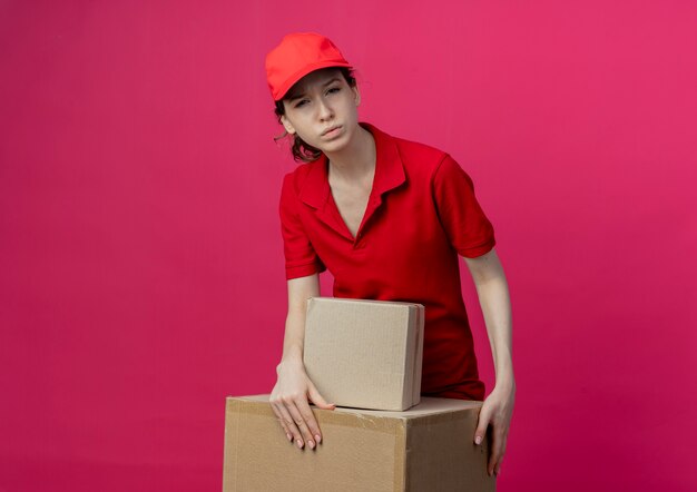 Premurosa ragazza di consegna graziosa giovane in uniforme rossa e cappuccio che mette le mani sulla scatola di cartone isolata su fondo cremisi con lo spazio della copia