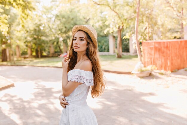 Premurosamente bionda giovane donna in abito romantico che guarda lontano mentre posa con alberi. Ritratto all'aperto di ragazza riccia triste in cappello di paglia retrò e abito bianco estivo.