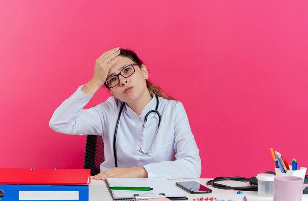 Premuroso giovane donna medico indossando abito medico e uno stetoscopio e occhiali seduto alla scrivania con strumenti medici guardando dritto mettendo la mano sulla testa isolata