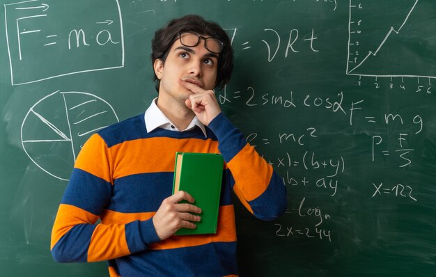 premuroso giovane insegnante di geometria con gli occhiali sulla fronte in piedi di fronte alla lavagna in classe tenendo il libro chiuso toccando il labbro guardando in alto