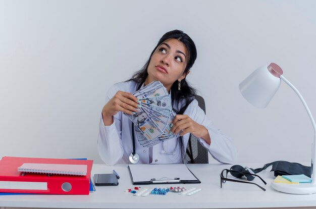 Premuroso giovane medico femminile che indossa abito medico e stetoscopio seduto alla scrivania con strumenti medici tenendo i soldi guardando il lato isolato