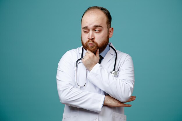 Premuroso giovane medico maschio che indossa cappotto medico e stetoscopio intorno al collo tenendo la mano sul mento e un'altra mano sotto il gomito guardando il lato isolato su sfondo blu