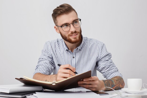 Premuroso manager maschio con occhiali rotondi, indossa una camicia formale, scrive su un taccuino mentre si siede al posto di lavoro,