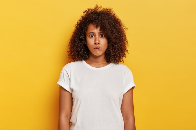 Preoccupata, adorabile ragazza con folti capelli ricci morde le labbra, ha un'espressione nervosa sorpresa, si sente imbarazzata a parlare in un luogo pubblico e indossa pose bianche su un muro giallo. Concetto di emozioni umane