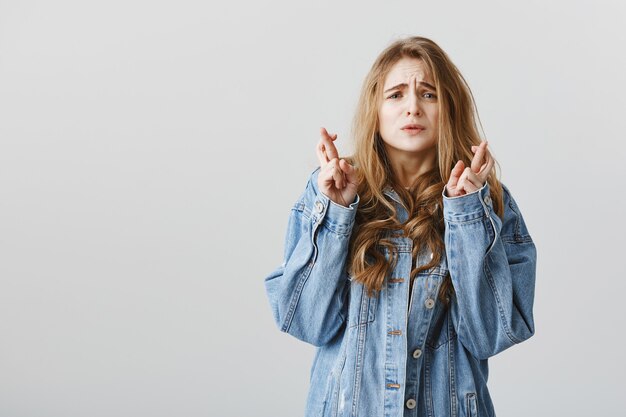 Preoccupata ragazza bionda speranzosa incrocia le dita, buona fortuna e aggrottando la fronte in attesa nervosa